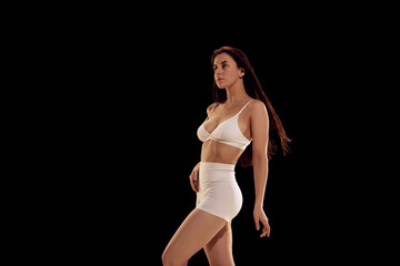 Fototapeta na wymiar Side view portrait of beautiful fit young woman in white sportwear posing with arms up against dark mode background. Concept of female health, fitness, sport, active lifestyle, dieting.