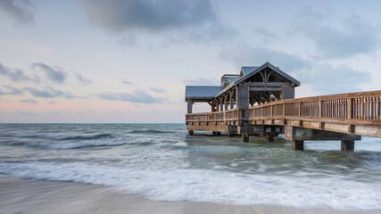 Wooden pier, with a covered open air structure at the end, reaching out into the tropical waters of the ocean, at sunrise. There is a hint of orange in the sky
