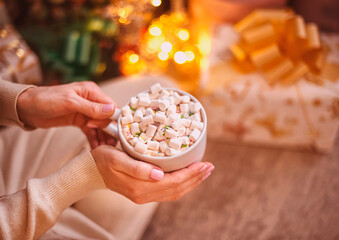 A white cup with cocoa and white marshmallows in female hands against a background of lights. Warm, cozy New Year's still life