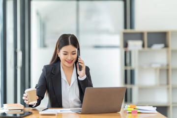 Fototapeta premium Shot of a young professional woman sitting at desk in front of laptop and using mobile phone working on business contract.