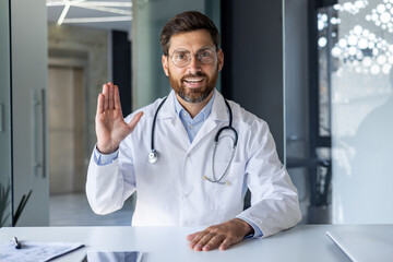 Portrait of a young male doctor in a white coat and with a stethoscope sitting at a table in a hospital, chatting online, greeting and waving at the camera