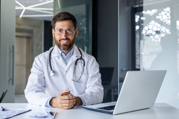 Portrait of a smiling young male doctor in a white coat sitting in the office of the clinic at the working table and confidently looking at the camera - obrazy, fototapety, plakaty