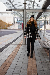 Woman in her mid-thirties with long red hair wearing a cap and black clothing in an urban location.