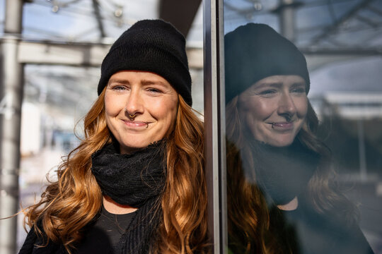 Woman in her mid-thirties with long red hair wearing a cap and black clothing in an urban location.