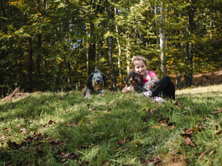 Naklejka na ściany i meble Happy little girl with two dogs sitting on a green grass lawn having fun, family picnics, spending time together, hiking in the mountains. People and pets outdoors lifestyle photo candid emotions.