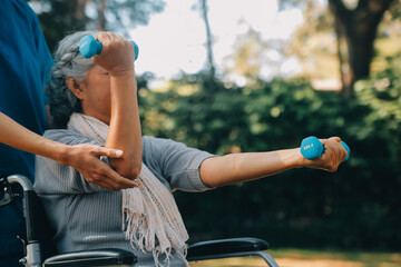Young nurse or physiotherapist in scrubs helping a happy retired old woman do fitness exercises with light weight dumbbells at home. Concept of physiotherapy for seniors - obrazy, fototapety, plakaty