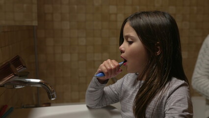 Daughter brushing teeth in front of bathroom mirror by sink while mother helps smaller brother to brush teeth, authentic family dental hygiene