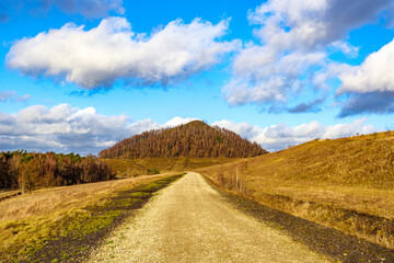 Hiking path between plain and hills with a mountain with autumn trees against the blue sky with white clouds in background, Thor Park - Hoge Kempen National Park, sunny afternoon in Genk, Belgium
