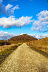 Fototapeta na wymiar Front view of dirt road between plains and hills, mountain with autumn trees against blue sky with white clouds in background, Thor Park - Hoge Kempen National Park, sunny afternoon in Genk, Belgium