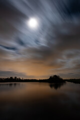 Scenic nighttime image of the moon over a lake while clouds pass by.