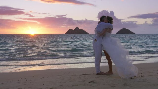Happy romantic couple in love getting married at cinematic sunset on Hawaii beach. Young man and woman holding hands kissing at beautiful sunrise. Love and romance, marriage, honeymoon holiday travel