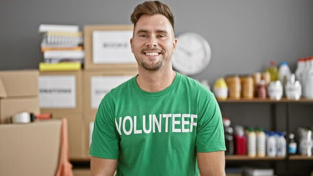 Handsome young man with beard wearing a 'volunteer' t-shirt standing in a warehouse full of boxes and supplies.