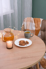  Kitchen home interior - Vitaminic healthy sea buckthorn tea in glass teapot with biscuits on white plate, cup of tea, burning candle, lights on wodeen table and chair with plaid.