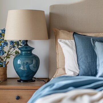 Close up of blue ceramic lamp on nightstand near bed with beige fabric headboard and blue pillows and blanket