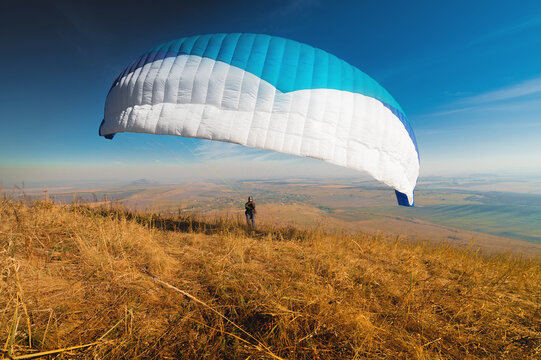 A paraglider takes off from a mountainside with a blue and white canopy and the sun behind. A paraglider is a silhouette. The glider is sharp, with little wing movement.
