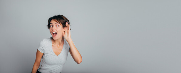 Banner photo of gorgeous female in casual wear listening to something by putting hand on the ear or try to hear you on distance and standing isolated on gray wall