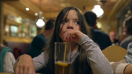 Pensive Little Girl Drinking Ice Tea with Straw at Restaurant in the Evening - Thoughtful Child Sipping Drink at Diner