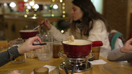 People eating cheese fondue at restaurant, closeup hand dips piece of bread into melted cheese at...