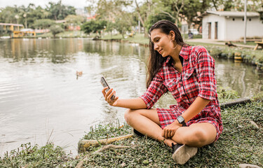 retrato de mujer joven sentada junto a un lago al aire libre y utilizando su teléfono móvil