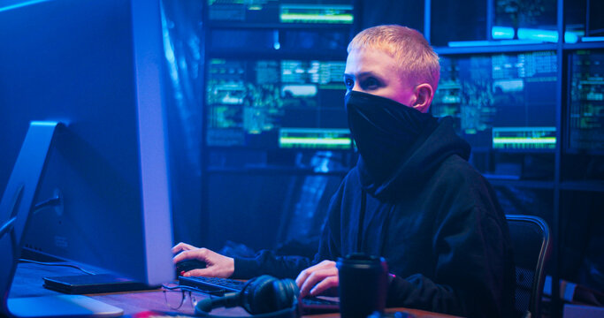 Caucasian woman hacker with blond short hair and masked face sitting in dark room with monitors and typing on keyboard of computer. Female hacking online system. Stealing information concept.
