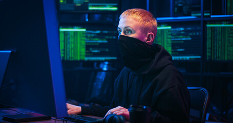 Caucasian woman hacker with blond short hair and masked face sitting in dark room with monitors and typing on keyboard of computer. Female hacking online system. Stealing information concept.