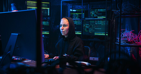 Caucasian joyful female hacker in hood sitting at table in technology monitoring room late in...