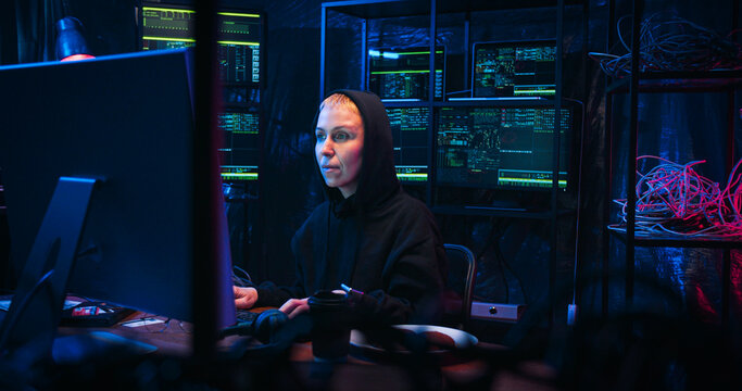 Caucasian joyful female hacker in hood sitting at table in technology monitoring room late in evening and working on computr. Woman hacking programs and spreading virus in internet.