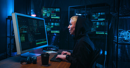 Side view on Caucasian female hacker in hood working on computer and typing in dark room. Woman...