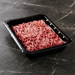 Raw and Fresh Minced Beef in Black Plastic Container just from the store, opened. On dark marble background. Minimalistic food photo, low-key.