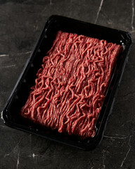 Raw and Fresh Minced Beef in Black Plastic Container just from the store, opened. On dark marble background. Minimalistic food photo, low-key.