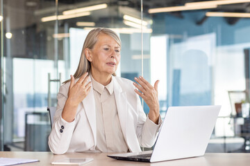 Serious senior woman works in the office, sits at a table with a laptop and waves his hands in his...