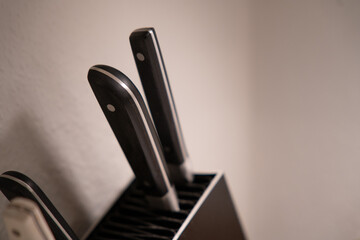 Black and stainless steel knifes in black knifeblock againt white wall, with dull lighting.