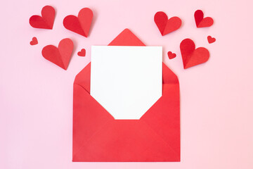 Red paper envelope with blank white note mockup inside and Valentines hearts on pink background. Flat lay, top view. Romantic love letter for Valentine's day concept.
