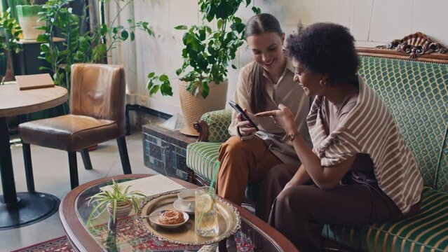 Medium shot of two cheerful multiethnic women, Caucasian and African American, sitting together on couch in cafe, looking at photos on smartphone, gossiping, commenting and laughingMedium shot of two 