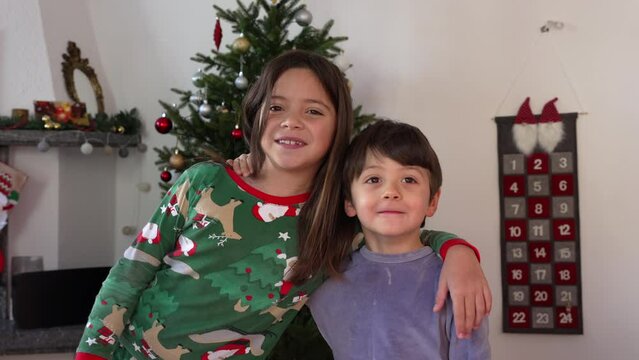 Siblings posing for camera with Christmas tree in background. sister and brother wearing pajamas smiling