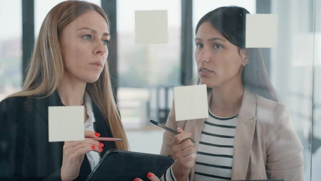 Video of two business young women working together on wall glass with post it stickers in the modern startup office