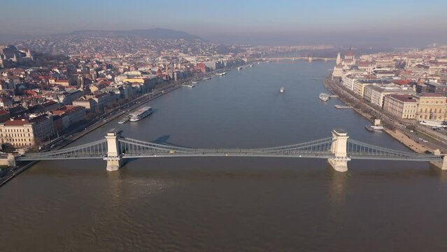 The morning sun illuminates the Chain Bridge and Budapest cityscape, with the serene Danube River flowing alongside. Slow Motion, Camera 4K RAW. 