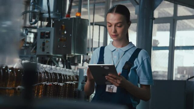 Medium stab footage of young Caucasian female manufacturing engineer standing with tablet near conveyor with moving soda bottles and typing data into laptop filling in protocol at beverage plantMedium