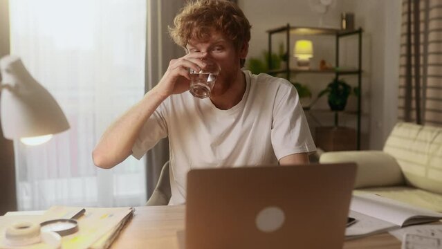 Handsome young man student or freelancer drinking clean water from glass while working on laptop computer typing browsing at home Busy male keep health balance hydration during day indoors