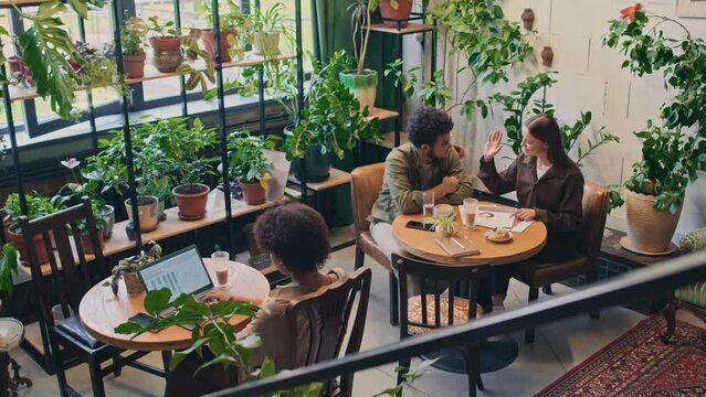 Full top shot of multiethnic customers sitting and doing business in stylish coworking space with lush greenery - Caucasian woman and Arab man discussing presentation, black woman typing on laptopFull
