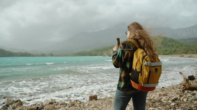 Turquoise sea water and waves against the backdrop of mountains. A rear view of a young woman, a travel blogger, taking photos for social media or as a keepsake. Strong winds have tousled her long hai