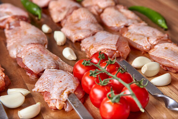 Metal skewers with raw uncooked chicken meat for frying on the wooden board with peppers near it, close-up perspective view. Shallow depth of field. Chicken and garlic in focus