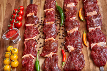 Metal skewers with raw uncooked beef meat for frying on the wooden board with peppers, spices and cherry tomatoes near it, close-up perspective view shallow depth of field. Meat and spices in focus