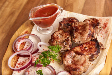 Part of the wooden board with fried beef meat with red onion rings, sauce and pita bread on the wooden table, close-up perspective view, shallow depth of field. Meat and onion in focus