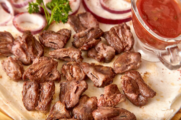 Part of the wooden board with small pieces of fried beef meat with red onion rings, sauce and pita bread on the wooden table, close-up perspective view, shallow depth of field. Meat in focus
