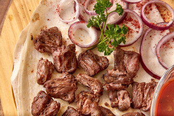 Part of the wooden board with small pieces of fried beef meat with onion rings, sauce and pita bread on the wooden table, close-up perspective view, shallow depth of field. Meat and onion in focus