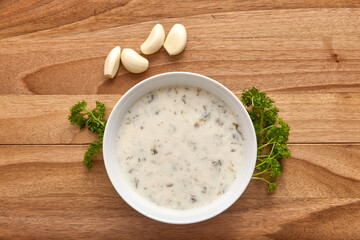 White plate with creamy soup on the wooden table with greens and garlic near it. Top view, flat lay