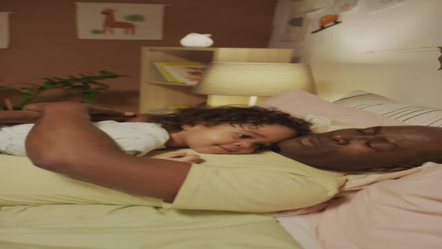 Vertical side slowmo of African American dad lying in bed in cozy decorated bedroom and hugging Biracial little toddler girl before sleep in eveningVertical side slowmo of African American dad lying i
