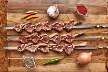Metal skewers with raw uncooked lamb meat for frying on the wooden board with chili peppers, garlic and onion, spices and salt near it, top view flatlay