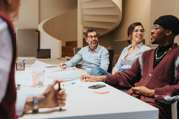 A multi-ethnic group of coworkers sits around a table during a business meeting, engaging in discussion with positive body language and expressions. - obrazy, fototapety, plakaty